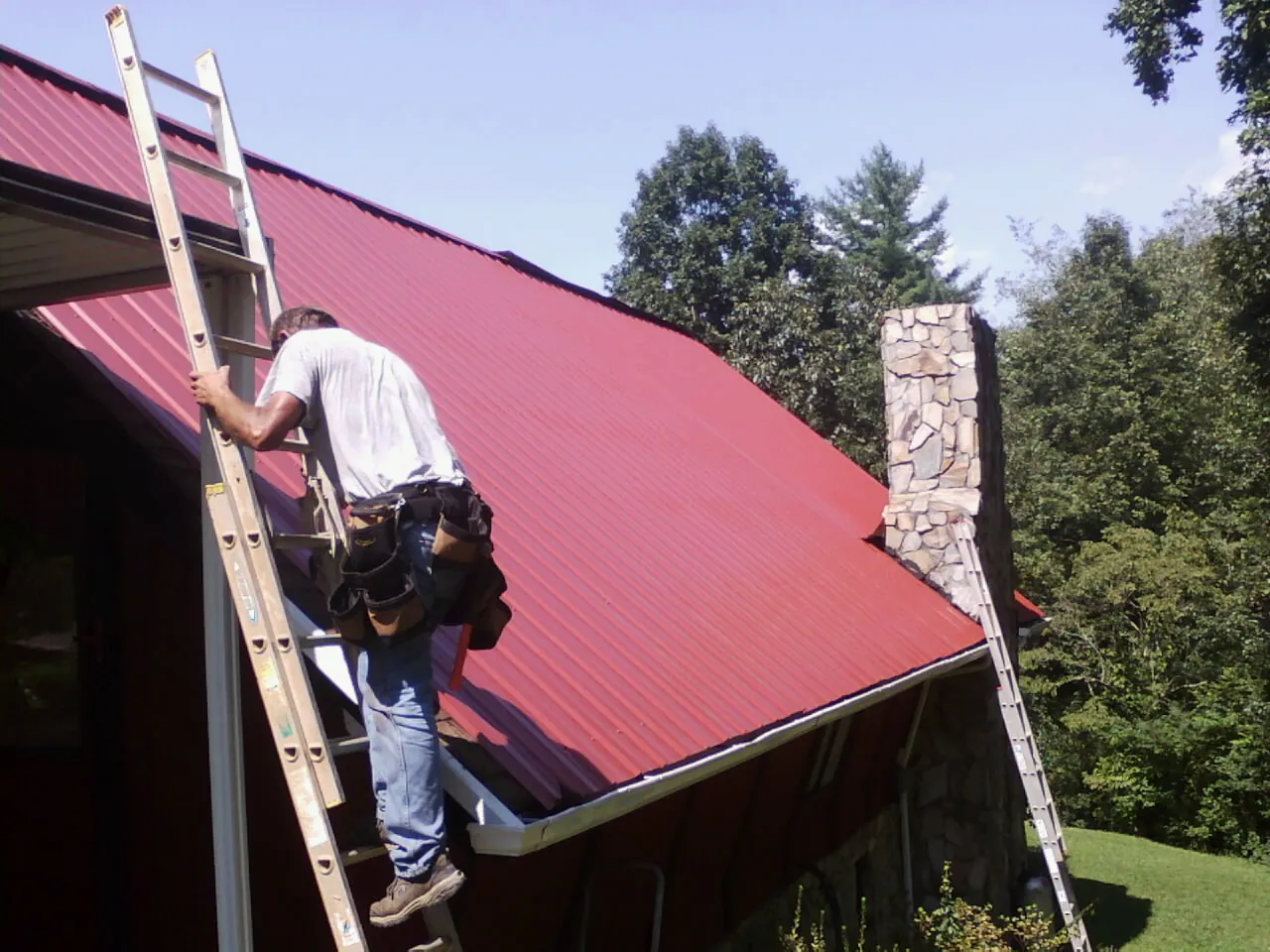 Roofer on a ladder beside a red metal roof and stone chimney — Soffit & Fascia Repair by Roofing Brothers in Culpeper
